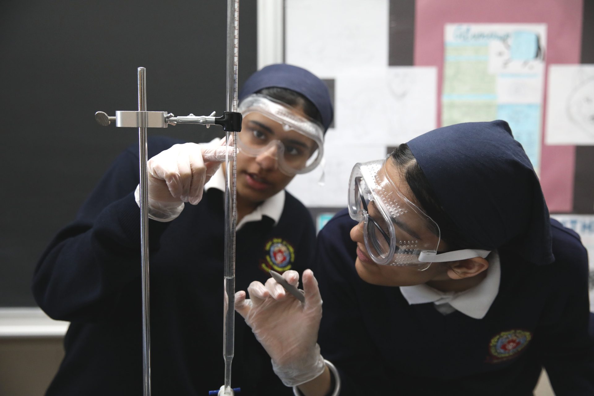 Two students in safety goggles and gloves reading a burette during a titration in the school science laboratory